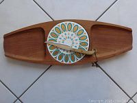 Wooden cheese platter with side compartments, mid-century ceramic plate, and ivory handled knife arranged on white tile floor.