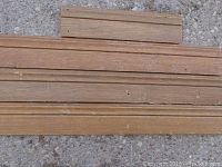 Close-up image showing several pieces of used mahogany baseboard trim laid out horizontally on concrete ground. Wood pieces are narrow and rectangular with visible grain and some paint marks or stains on edges and surfaces.