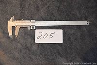 Photo showing a full-length metal precision caliper resting on a dark fabric surface alongside a card labeled 205