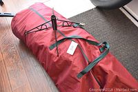 Red fabric bag with green handles containing the artificial Christmas tree and a black metal tree stand placed on a hardwood floor.