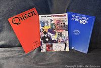 Three piano music books displayed: Queen, Elton John, and Song Classics of the 60's - showing front covers.