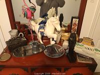 Wide angle of dresser top showing multiple decor items including the white horse figurine, metal flamingo, silverplated cup and tray, ceramic vases, metal box and Lenox plate.