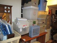 Photo showing multiple stacked clear plastic storage containers with white lids, alongside blue plastic bins and a blue crate.