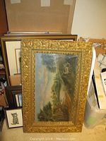 Stacked framed artworks including gilt landscape painting with deer and other paintings behind