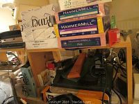 Wooden shelf with multiple packs of colored Hammermill paper and various office supplies including a hole punch and laminator.