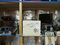 Shelf view showing assorted cameras in cardboard box labeled "Camera/Slide Stuff", brass spyglass and camera bag