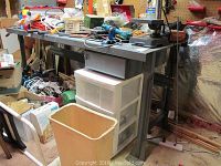 Full view of the metal work table with various tools and objects on the tabletop, a waste bin in front, and storage under the table in a basement or workshop setting.