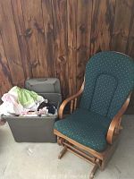 Wooden rocking chair with green patterned cushions next to grey tote bin filled with fabric and blankets, against wood-paneled wall on cement floor.