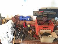 Photo of various hand tools arranged on a workbench, including hammers, screwdrivers, pliers, wrenches, and a work belt with a blue flashlight and a red power sander nearby.