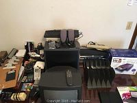 Overview of computer monitor, HP printer, keyboard, speakers, small black clipboard, and assorted small electronics and office supplies on table.