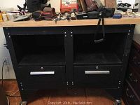 Front view of the black workbench showing two open shelves and two drawers with metal handles, topped with various items which are not included.