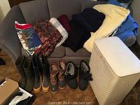 Full view of four pairs of men's boots alongside multiple folded blankets and a laundry hamper on a couch and floor.