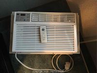 GE window air conditioning unit with power cord and front grille with control panel and remote on top.