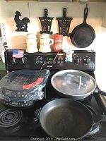 View of stove top with cast iron pots, skillet, kitchen decor items, and the induction cooktop sealed in plastic on stove.
