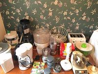 Overview of various kitchen appliances on wooden surface against floral wallpaper background. Items include food dehydrator, toaster, blender base, juicer parts, coffee pot, and accessories.