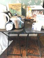 Two black folding tables supporting kitchen appliances and serveware including pressure cooker, electric kettle, hand mixer, pizzelle baker box, and six goblets.