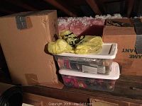 Boxes of Christmas decor including ornaments in clear plastic bins and cardboard boxes on attic floor with exposed insulation and wooden beams