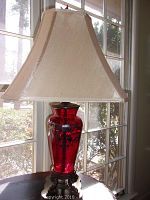 Full view of lamp on table near a window showing the beige square bell-shaped fabric shade and red glass body with black floral detailing.