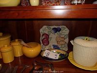 Shelving display showing three yellow ceramic canisters with lids, yellow mortar and pestle, decorative plates with fruit motif, white hobnail covered dish with yellow base.