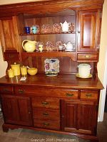 Wooden dining room hutch with upper display cabinet containing glass shelf and ceramic dishware, and lower base with drawers and cupboards, viewed from the front.