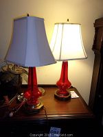 Pair of red glass lamps with white fabric shades on a wooden table, both switched on and working