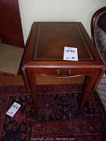 Front view of the wooden side table showing the leather-like top with gold border and drawer with handle.