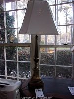 Front view of the brass buffet lamp placed on a dark wooden surface in front of a window showing the full height and lampshade.