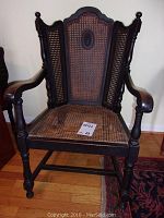 Front-facing view of the black painted wood chair with cane seat and back showcasing design and wear on arms.