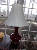 Front view of purple etched glass lamp with white bell-shaped shade on wooden table in front of window. Lamp base shows brass or bronze finish with cord visible.