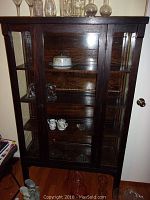Full view of the dark wood display cabinet showing front glass doors and internal shelves with a few glass and porcelain items on shelves and floor around it