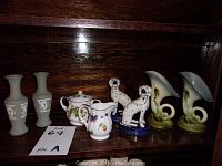 Full view of all ceramic figurines and vases, arranged on a wooden shelf in single row, showing variety including cameo vases, dog figurines, floral teapot and creamer, cornucopia vases.
