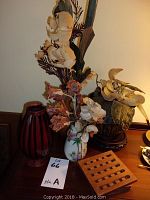Four decorative items on table including two floral arrangements, a red vase and a wooden trivet.