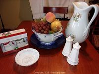 Full view of all six items on table including fruit bowl with artificial fruit, large floral pitcher, salt and pepper shakers, small decorative plate, and boxed holiday salt and pepper set.