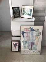 Four framed artworks displayed with three watercolors and one large poster leaning against a dresser and wall.