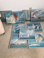 10 framed nautical wall art prints featuring sailboats with colorful sails on water and blue skies, displayed in a collage on the floor.