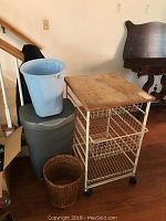 Kitchen cart with butcher block top, white wire shelf frame, and caster wheels; one wicker and two plastic wastebaskets next to it