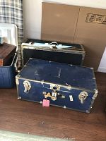 Two foot lockers, one navy blue in front with metal edges and worn exterior, the other dark colored behind. Both have handles and latches.