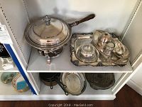 Silverplate chafing dish with lid, silverplate tray holding gravy boat and serving dishes on white shelving.