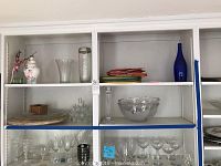 Four white shelves with assorted clear glassware including vases, bowls, candlesticks, colorful stacked plates, and glassware like wine glasses.
