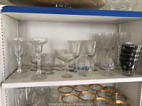 Four shelves displaying assorted glassware: clear and gold-rimmed fruit cups, various crystal stemware including water and champagne glasses