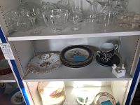 Shelf with clear glassware including wine glasses, cocktail glasses, bowls, and small dishes. Below, shelves hold various china plates and a cup and saucer set.