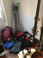 Photo showing skis with bindings, ski poles, assorted gloves, mittens, boots and maroon backpack arranged on floor against wall.