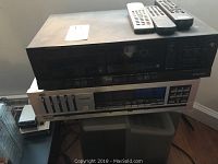 Kenwood stereo equipment stacked with another audio device, remote controls on top, cords visible, showing front panels with buttons, dials and display.
