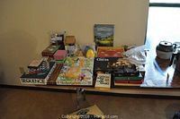 Full table view of the games lot showing all boxed games and containers arranged on a folding table with beige wall background.