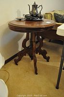 Full view of round wooden pedestal side table with silver tea set on top, tabletop attached.