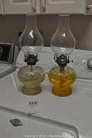 Pair of matching glass oil lamps on a laundry machine, showing honeycomb patterned bases filled with oil and clear glass chimneys.
