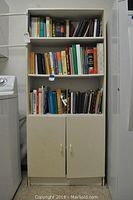 White shelving unit with three shelves filled with cookbooks of various colors and wear. Cabinet doors below are closed.