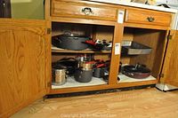 Open wooden cabinet displaying nonstick and stainless steel pots, pans, and baking pans with lids and red handles.