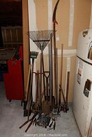 Wide view showing 3 rakes, 3 tall shovels, 3 axes, pick, and various garden hand tools leaning against a wall.