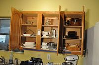 Wide view of oak kitchen cabinet with doors open showing plates, bowls, mugs, glassware, and storage containers, with KitchenAid mixer in foreground.